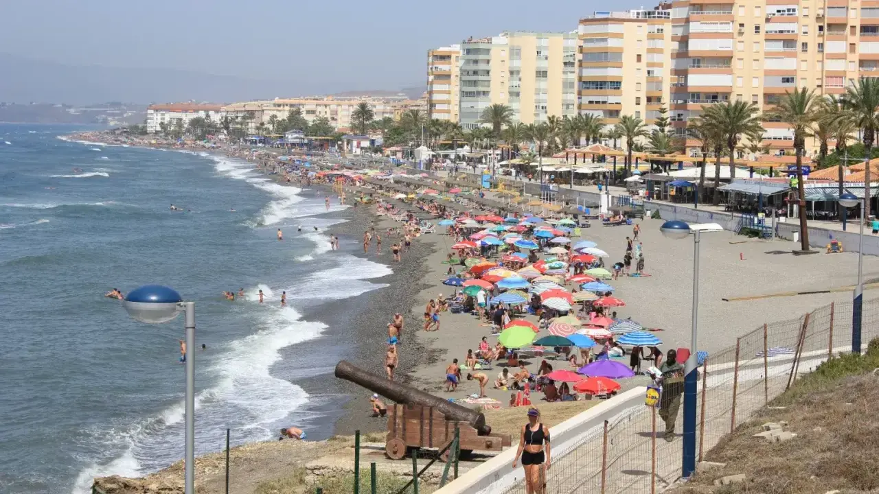 Sonniger Torrox Strand mit vielen bunten Sonnenschirmen, Menschen im Meer und an Land, sowie historischen Kanonen.