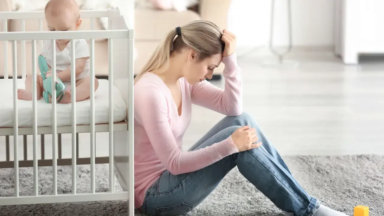 Zmartwiona mama siedzi obok łóżeczka, zastanawiając się, depresja poporodowa ile trwa. Dziecko bawi się w łóżeczku.