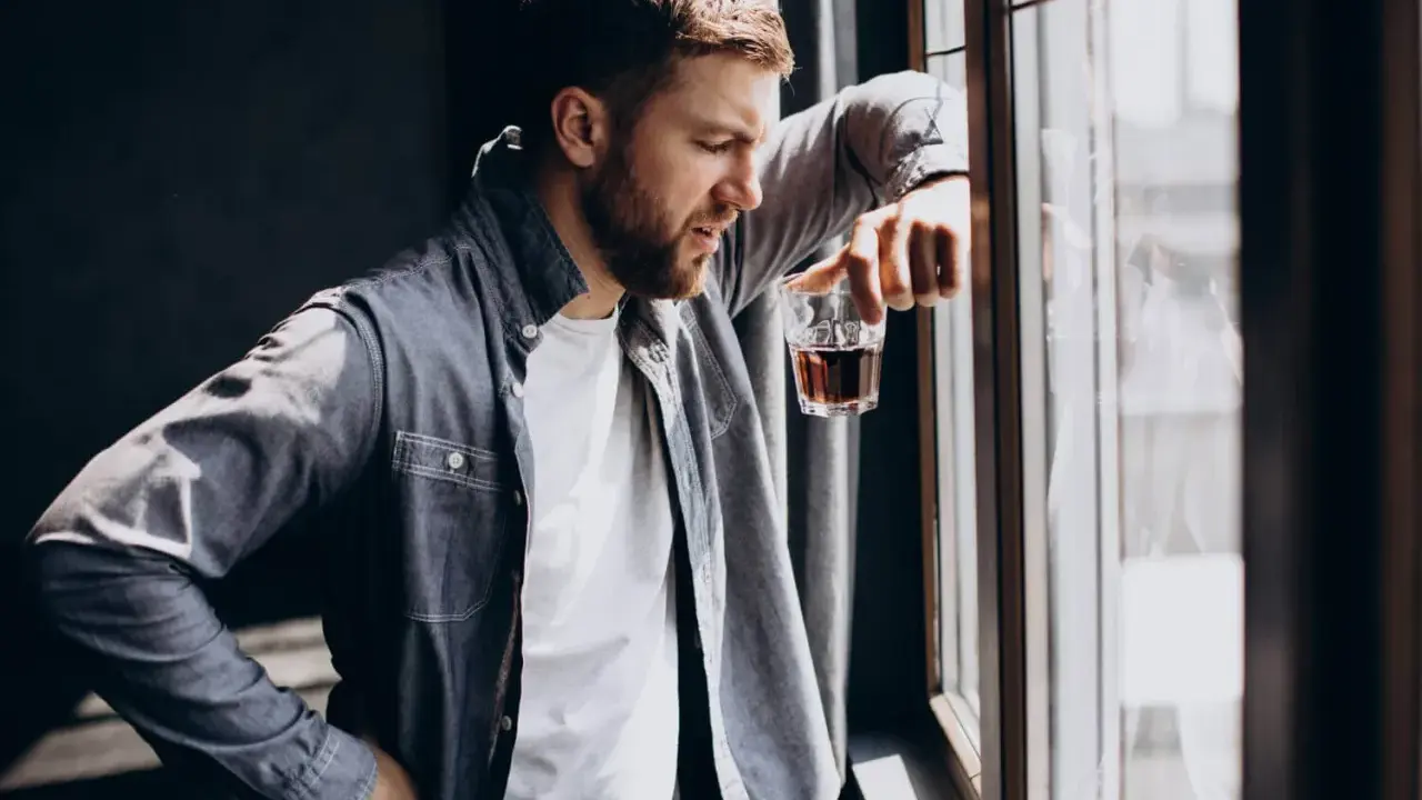 Mężczyzna zamyślony przy oknie, trzymający szklankę z bursztynowym płynem. Czy to jeden z ukryty alkoholizm objawy?