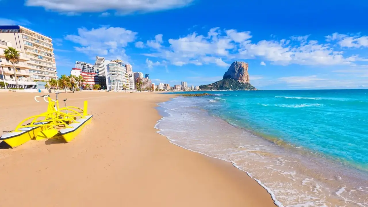 Sonne, Strand und Meer in Calpe, Spanien. Ein gelbes Wasserfahrrad wartet am Sandstrand, im Hintergrund die Stadt und der markante Felsen Peñón de Ifach. Ein echter Geheimtipp!