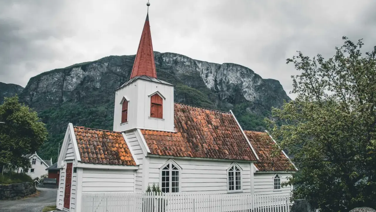 Biały kościółek z czerwoną wieżą i dachem, otoczony białym płotkiem, na tle surowych gór. Symbol norweskiej religii i spokoju.