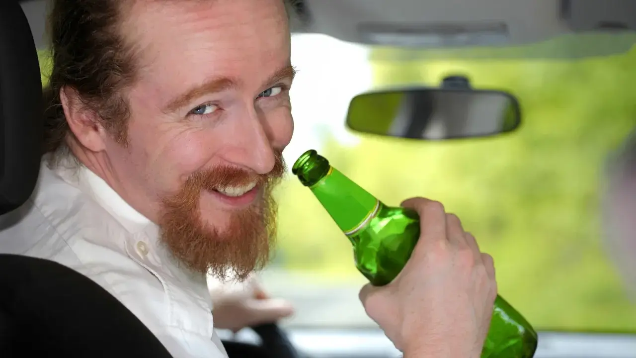 Uśmiechnięty mężczyzna z rudą brodą trzyma zieloną butelkę piwa w samochodzie. Mówi, że piwo to nie alkohol, ale jazda z nim jest niebezpieczna.