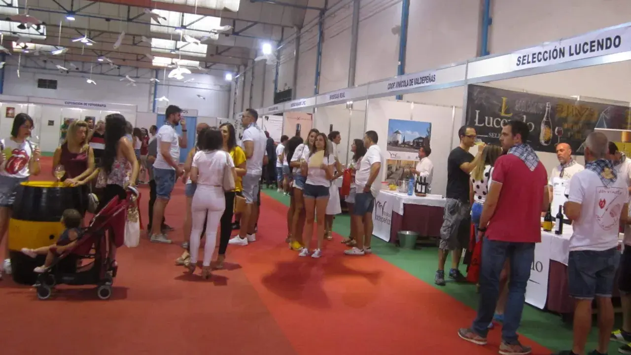 Gente en un evento de degustación de vinos, con stands de bodegas como "Coop Vinícola de Valdepeñas". El "túnel del vino Valdepeñas" se siente en el ambiente.