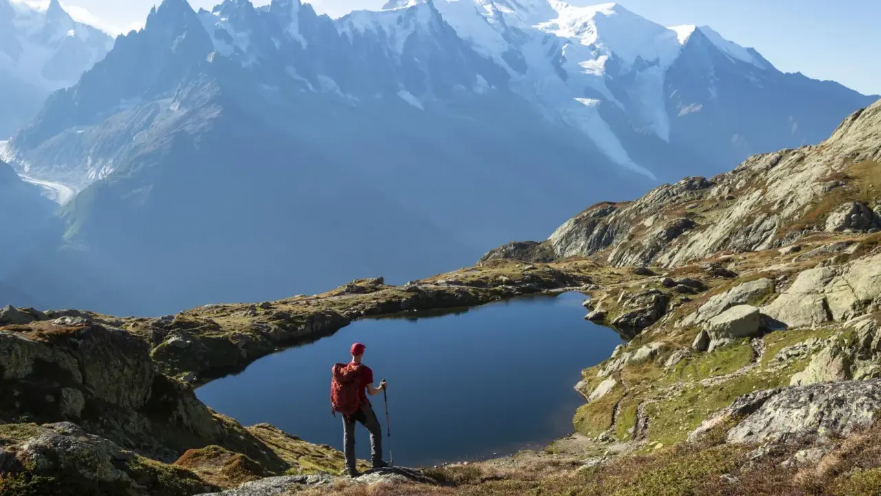 Widok na wejście na Mont Blanc. Turysta z plecakiem i kijkami stoi nad górskim jeziorem, podziwiając ośnieżone szczyty.