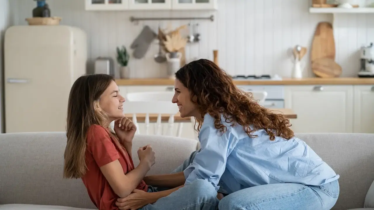 Madre e hija ríen en el sofá. Ella le enseña como es la primera regla de una niña: la empatía.