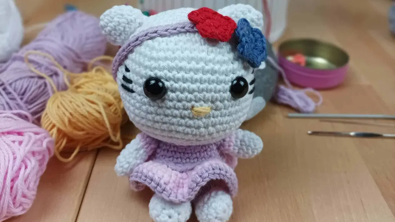 Muñeca de Hello Kitty tejida a crochet, con vestido lila y lazo rojo y azul. Hilos y agujas de crochet alrededor, perfectos para crear tus propios hello kitty crochet patrones español.