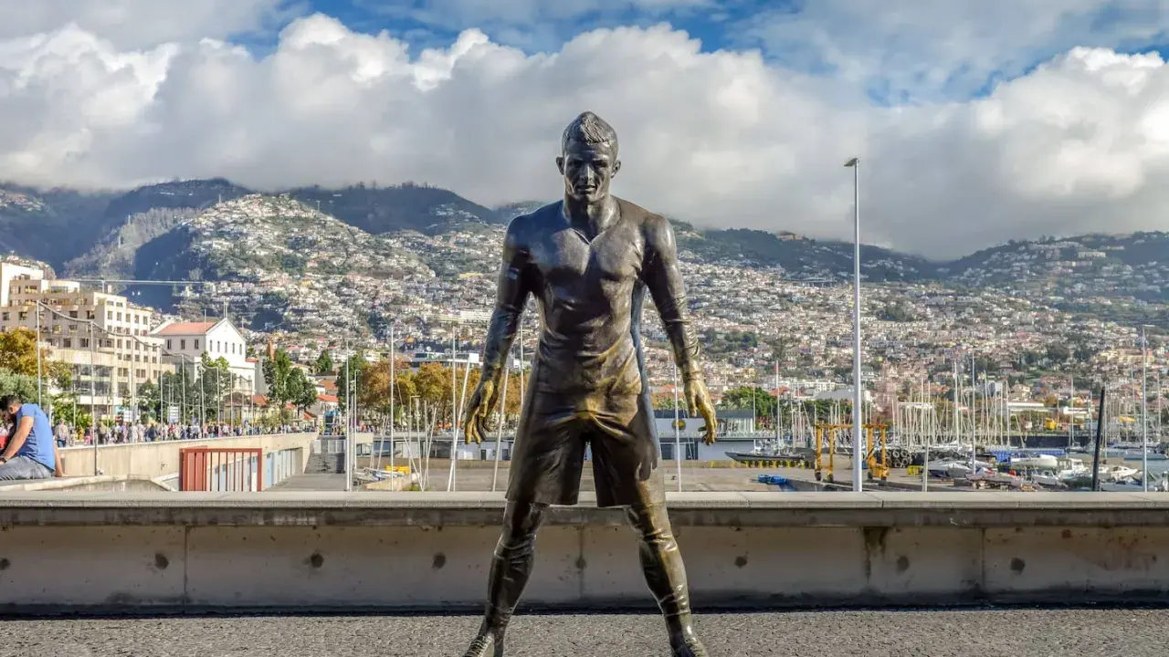 Bronzestatue von Cristiano Ronaldo am Hafen von Funchal, Madeira. Hinter ihm erstreckt sich die Stadt bis zu den Bergen. Ein Denkmal für den weltberühmten Fußballer, das man fast wie am **cristiano ronaldo flughafen** erwarten könnte.