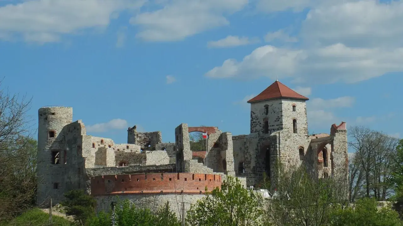 Zamek Tenczyn, ruiny kamiennej twierdzy z czerwoną cegłą i polską flagą, na tle błękitnego nieba z chmurami.