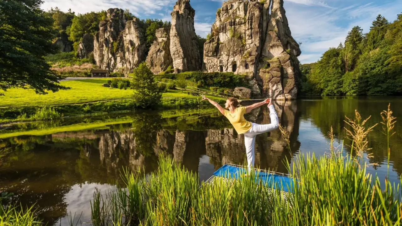Frau praktiziert Yoga am See vor den Externsteinen NRW. Die Felsen spiegeln sich im Wasser.