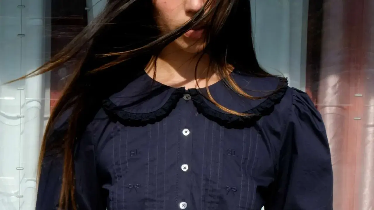 A young woman with windswept hair wears a dark blue blouse with a charming peter pan neckline, delicate embroidery, and puffed sleeves.
