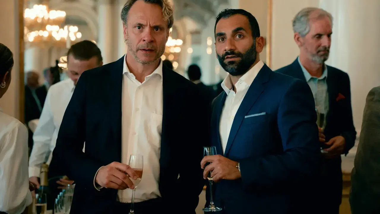 Två män i kostym håller glas med champagne. Bakom dem syns en festlig miljö med ljuskronor. Kanske en scen från "hundarna" på SVT.