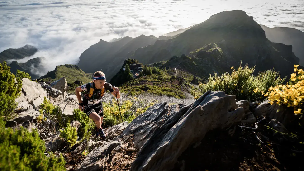 Läufer kämpft sich auf Madeira bergauf, vorbei an Felsen und gelben Ginsterbüschen, mit Wolkenmeeren und schroffen Gipfeln im Hintergrund.