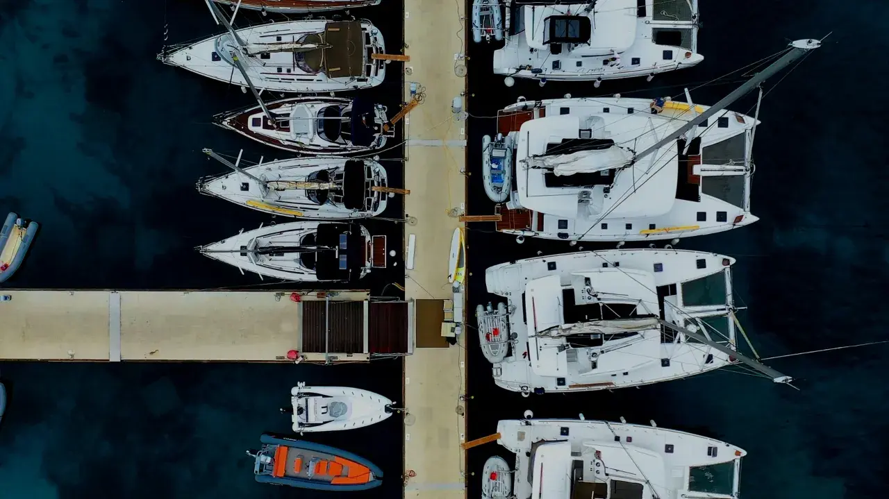 Vue aérienne d'un port avec plusieurs yachts à voile blancs amarrés, certains catamarans, et des annexes gonflables.