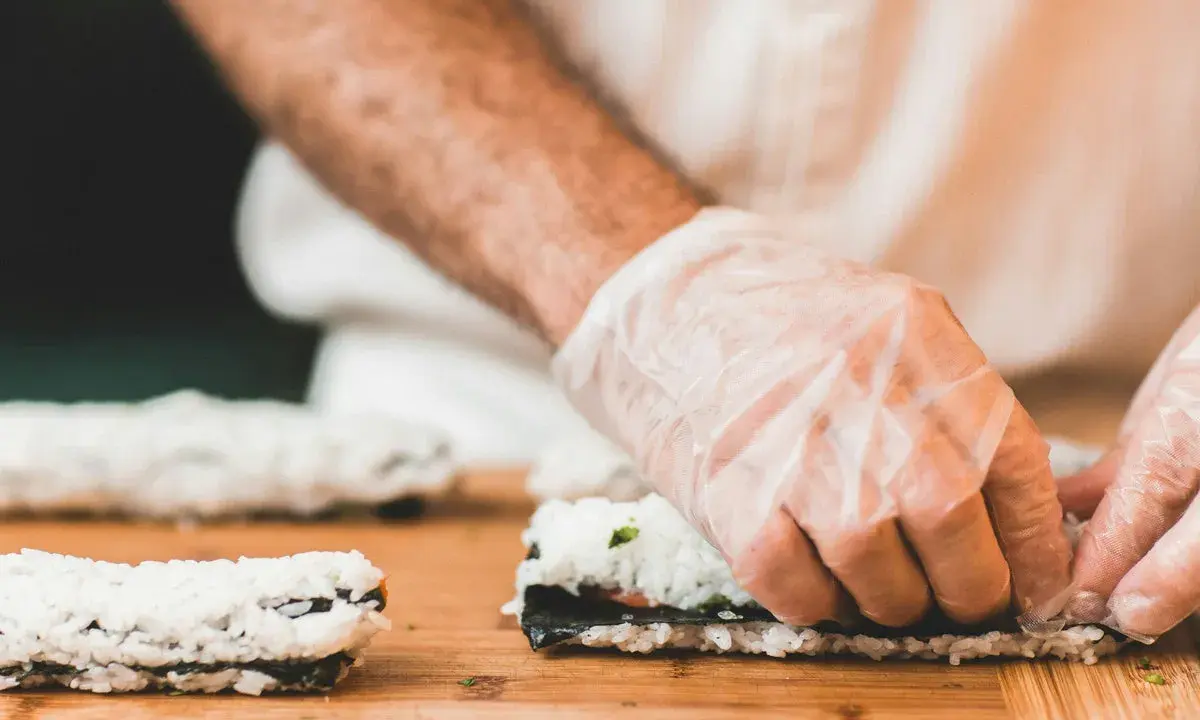 Hände mit Handschuhen rollen Sushi auf einem Holzbrett. So einfach ist Sushi selbst machen!
