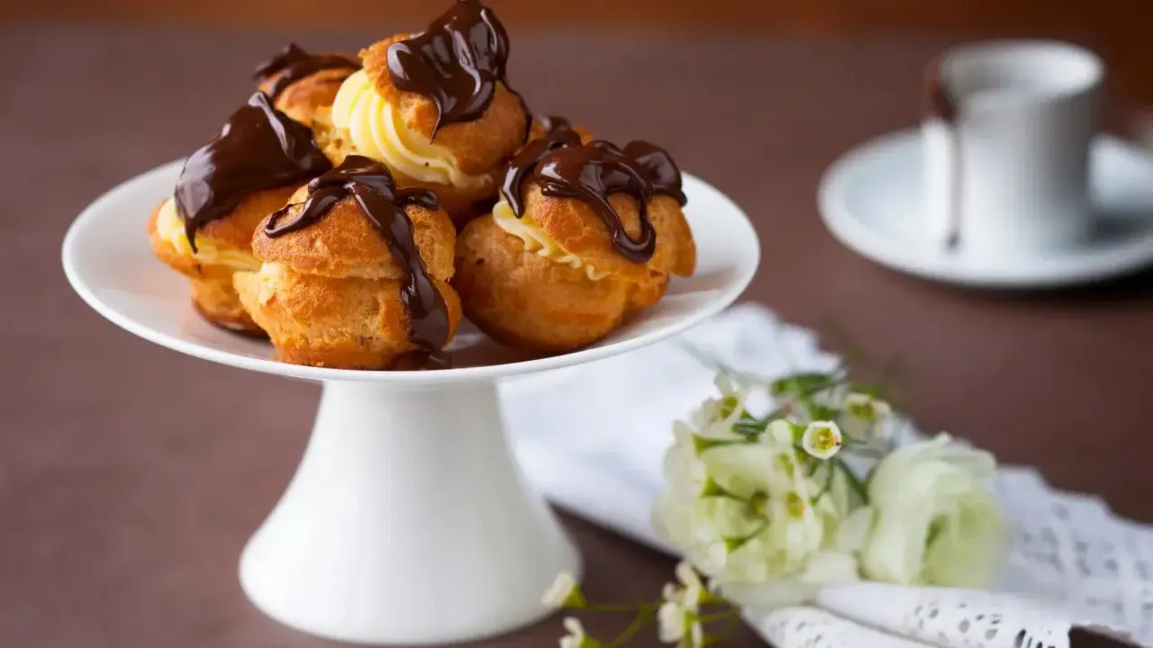 Un plateau blanc présente des profiteroles gastronomiques garnies de crème et nappées de chocolat noir. Des fleurs délicates ajoutent une touche de fraîcheur.