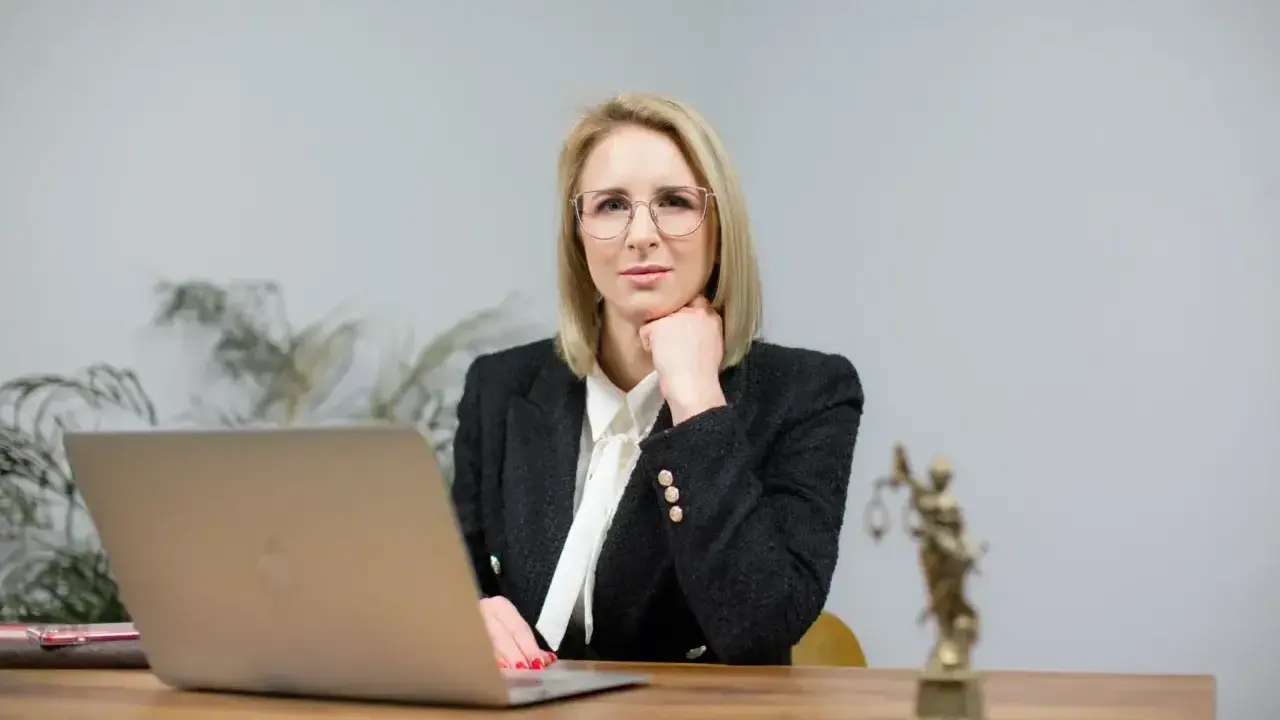 Kobieta w okularach i żakiecie siedzi przy biurku z laptopem i Statuą Temidy.