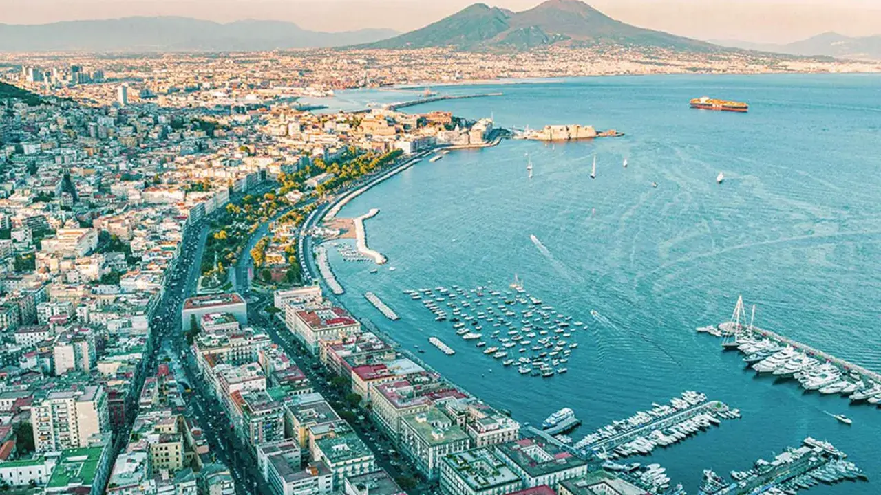 Vy över Neapel med Vesuvius i bakgrunden. Hamnen är full av båtar, och staden sträcker sig längs kusten. En perfekt vy innan man flyger från neapel flygplats.