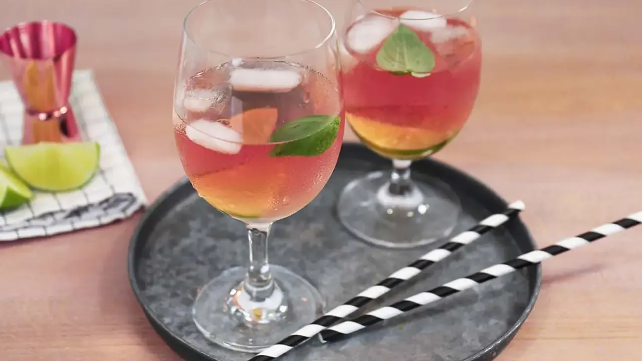 Zwei Gläser Rosato Spritz mit Eis und Minze, dazu Limettenspalten und ein Messbecher. Perfekt für einen sommerlichen Genuss.