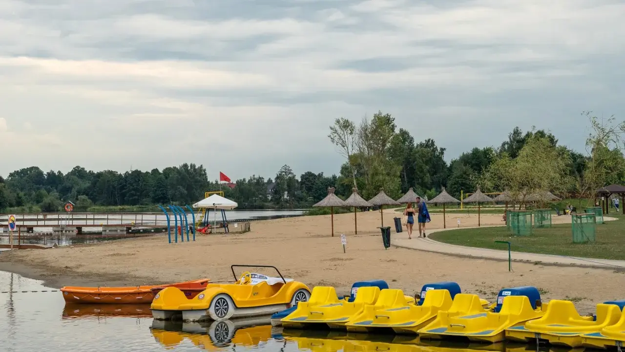 Żółte rowerki wodne i łódki czekają na plaży nad jeziorem blisko Warszawy. Para spaceruje po piasku pod słomianymi parasolami.