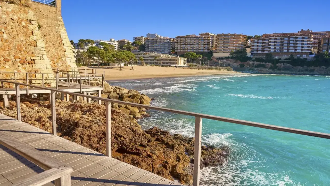 Paseo marítimo de madera junto a rocas y mar turquesa, con playa y edificios al fondo. Un tramo del camí de ronda Tarragona.
