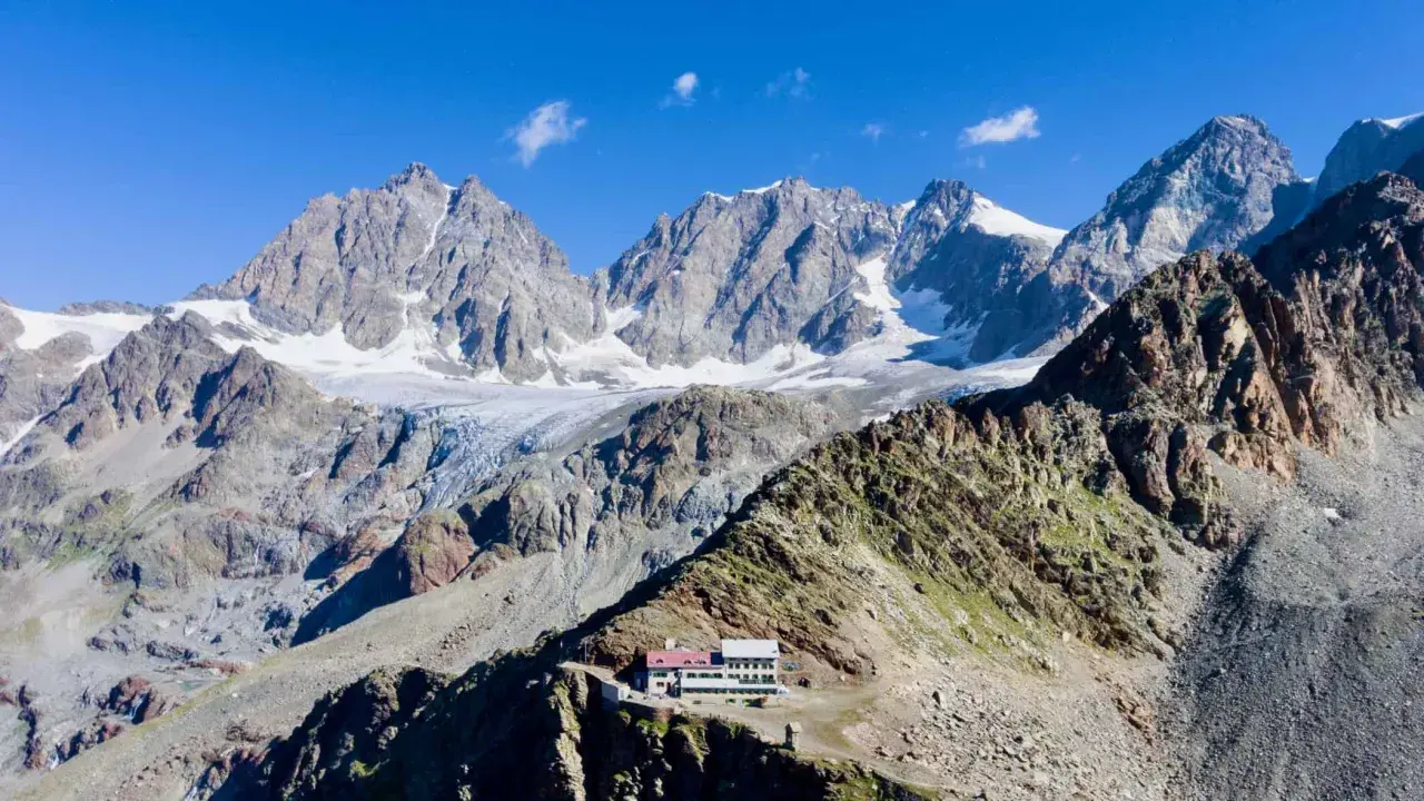 Il rifugio Marinelli, un edificio bianco e rosso, si erge su una cresta rocciosa con imponenti montagne innevate sullo sfondo.