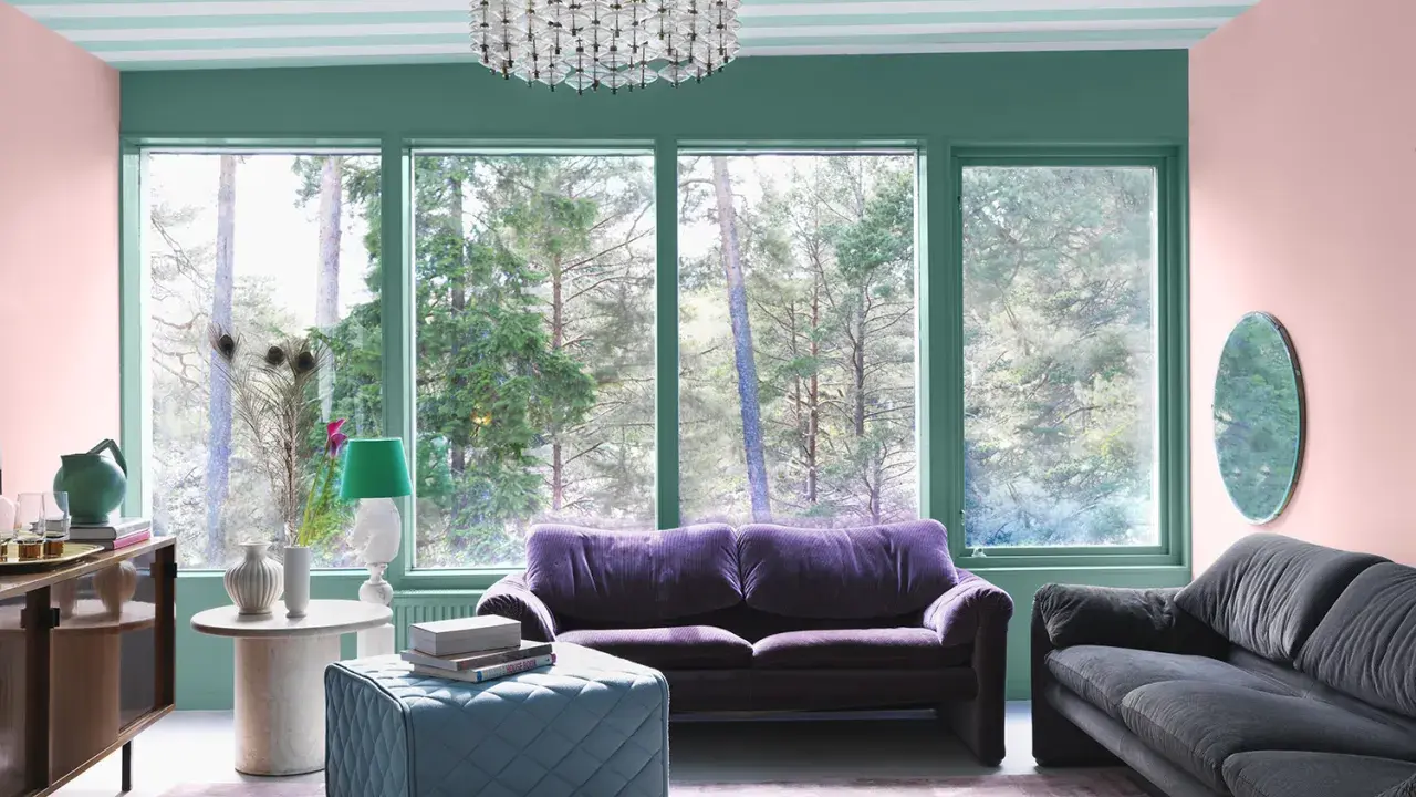 Ett vardagsrum med art deco inredning, mjuklila soffa, mörkgrå soffa, turkos pall och en stor fönsterutsikt över skogen.