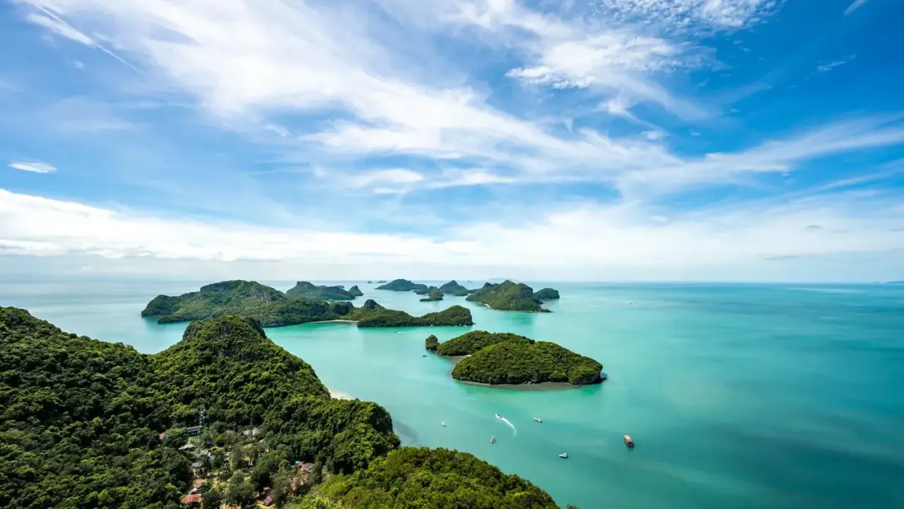 Grüne Inseln ragen aus türkisfarbenem Wasser im Nationalpark Thailand. Kleine Boote gleiten über die ruhige See unter einem blauen Himmel mit weißen Wolken.
