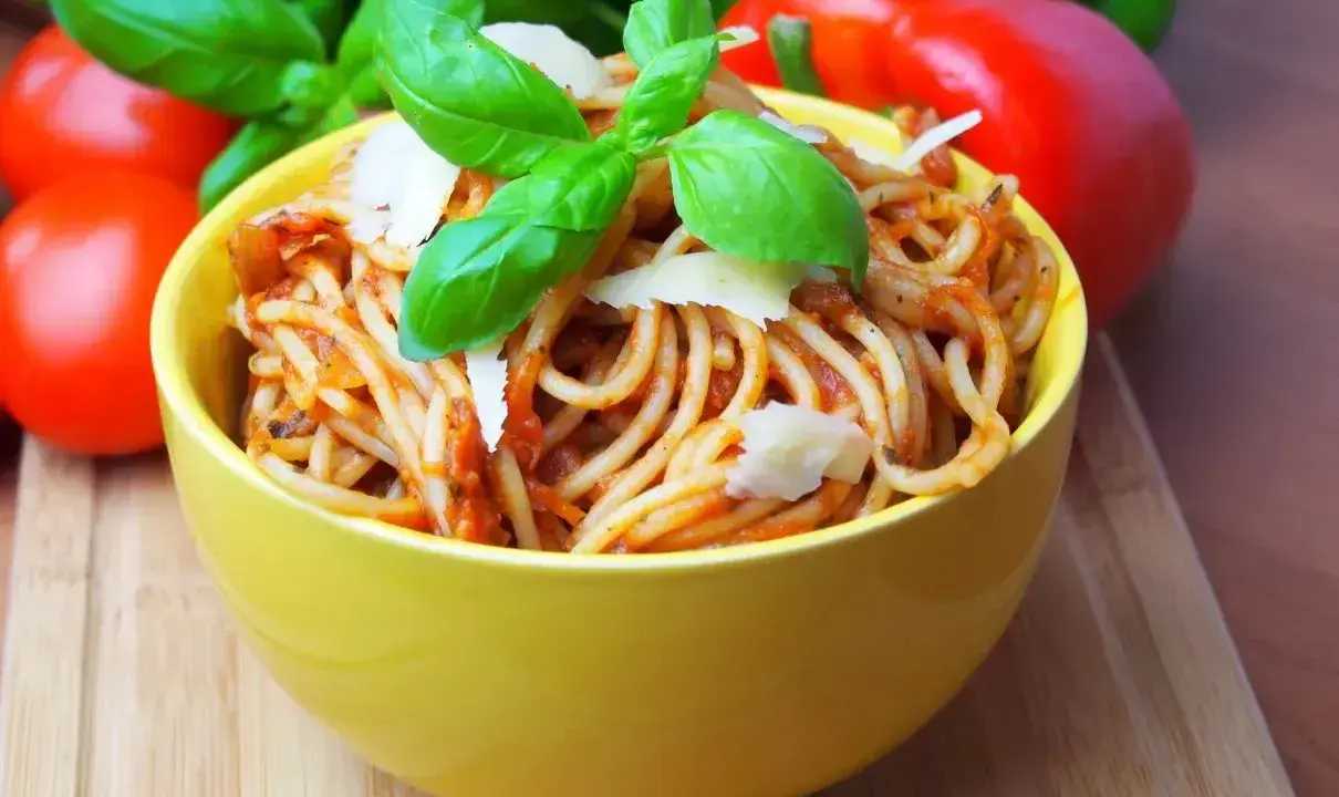 Pyszne spaghetti bez mięsa w żółtej misce, udekorowane bazylią i parmezanem. Obok pomidory i papryka.