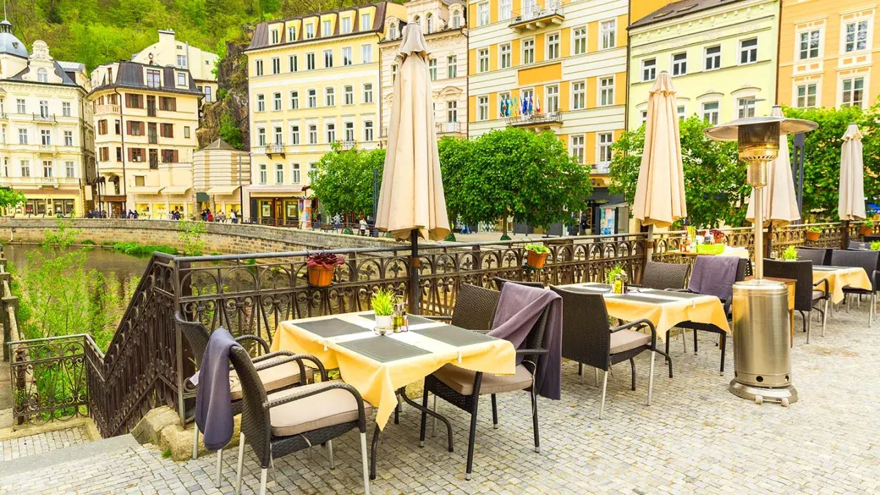 Uteservering vid en flod i Karlovy Vary med gula byggnader i bakgrunden.
