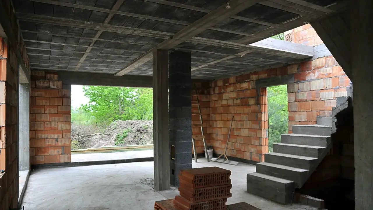 Wnętrze budowy z widocznym stropem gęstożebrowym, betonowymi schodami i ceglanymi ścianami.