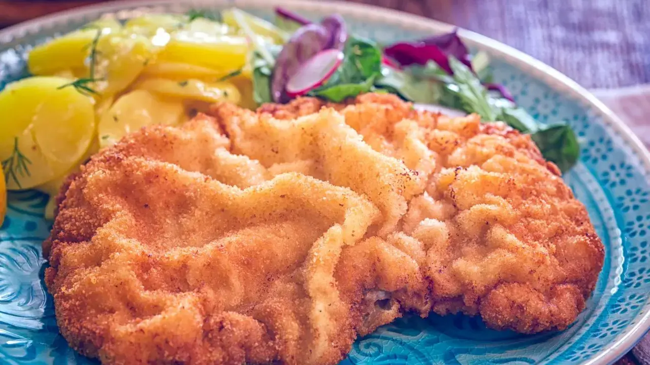 Ein knuspriges, originales Wiener Schnitzel auf einem blauen Teller, serviert mit Kartoffelsalat und frischem Grün.
