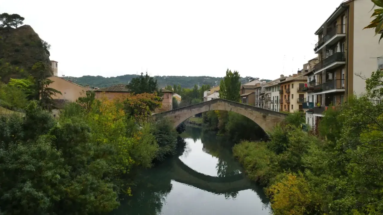 El puente de la cárcel de Estella cruza un río sereno, reflejando su arco de piedra entre frondosa vegetación y edificios históricos.
