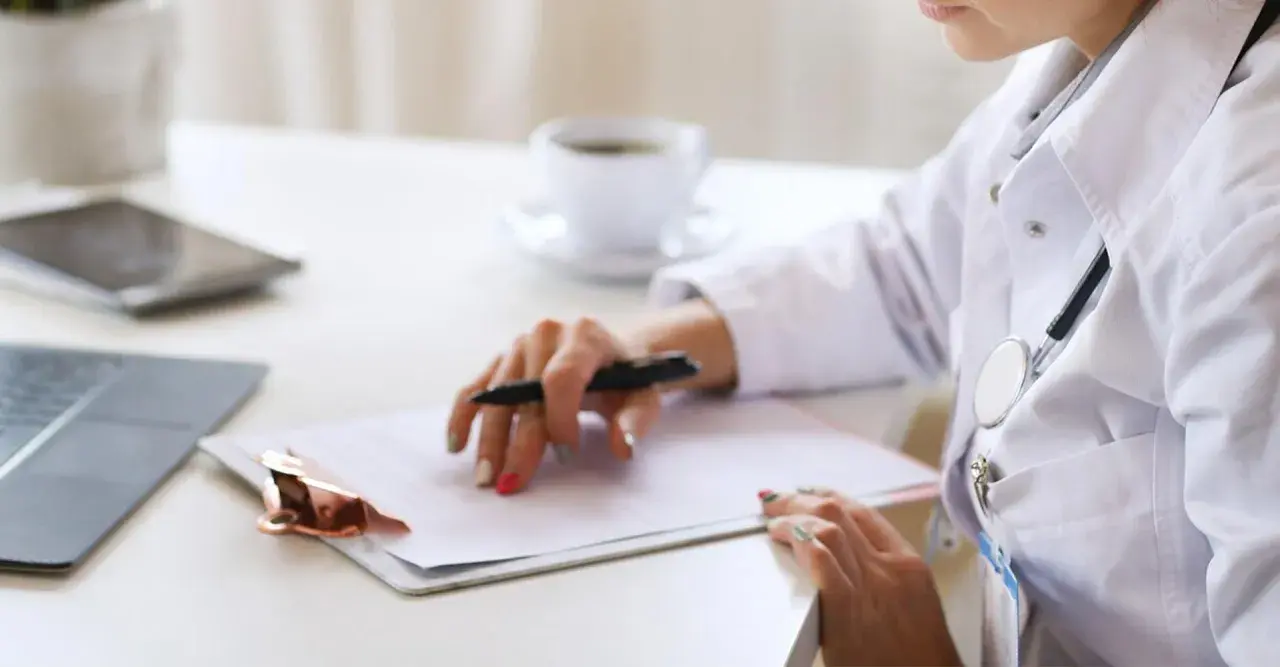 Médica revisa historial para tratamiento de hiperplasia endometrial. En su escritorio, laptop, tablet y café.