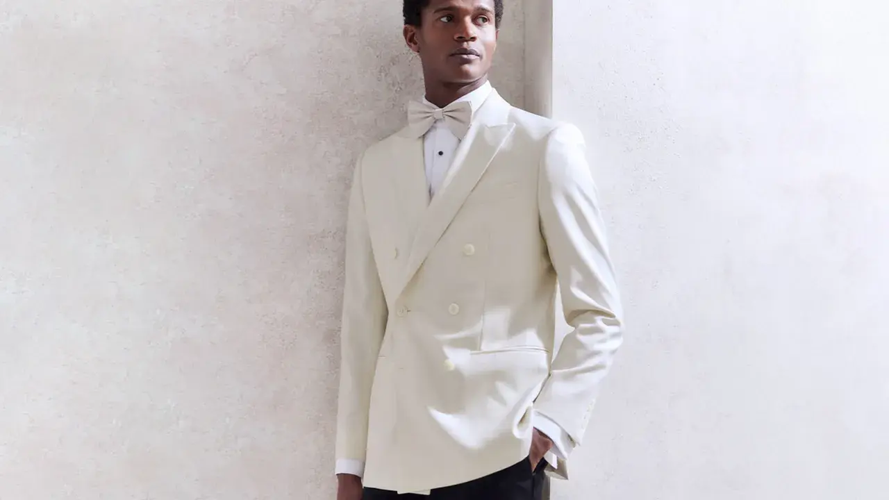 A Black tie wedding guest man in a cream double-breasted tuxedo with a matching bow tie, looking dapper.