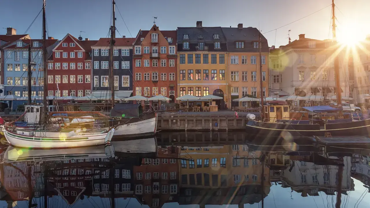 Färgglada hus vid en kanal i Köpenhamn, med båtar i förgrunden. Solen skiner och reflekteras i vattnet. En perfekt vy för en flyg till Köpenhamn-resa.