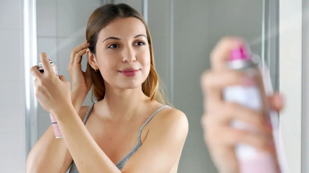 Frau sprüht sich Trockenshampoo ins Haar, um es aufzufrischen. So einfach ist es, wie man Trockenshampoo benutzt.