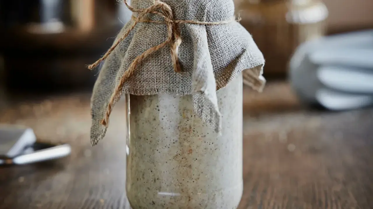 En burk med bubblig surdegsstarter, täckt med tyg och snöre, står på ett träbord.