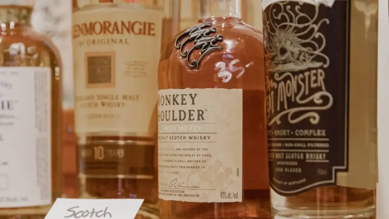 Flera flaskor whisky, inklusive Monkey Shoulder och Glenmorangie, står uppradade. En liten skylt med texten "Scotch" syns framför.