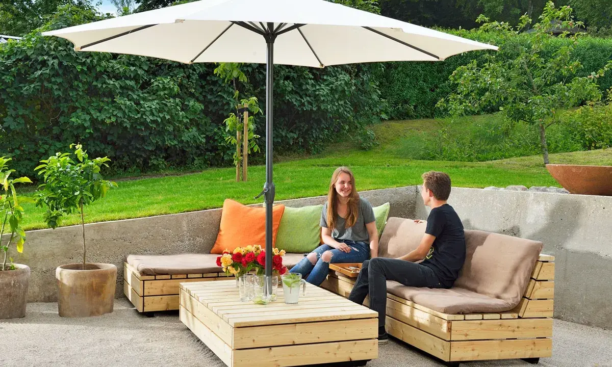 Två personer njuter av solen under ett parasoll vid en hemmabyggd loungesoffa i trä.