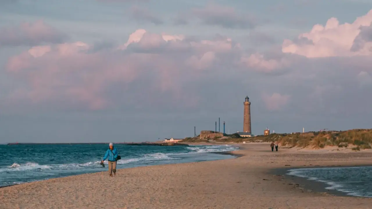 En person promenerar på stranden vid Skagens fyr. Molnen färgas rosa i solnedgången.