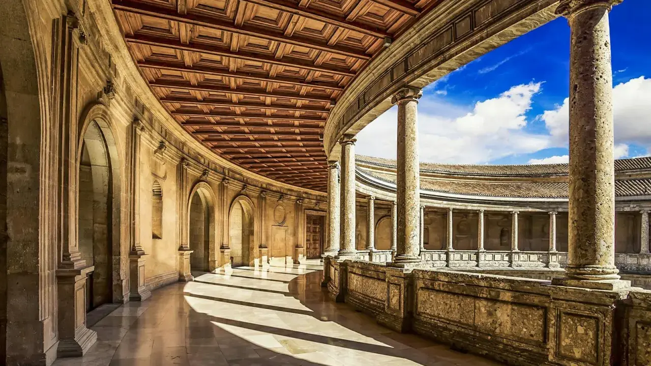 Ein sonnenbeschienener Arkadengang mit kunstvoller Holzdecke und Säulen, der zum Innenhof der Alhambra Moschee führt.