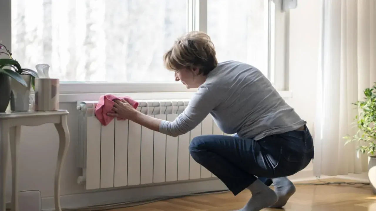 Mujer limpiando un radiador blanco con un paño rosa. Un ejemplo de cómo limpiar radiadores para mantener el hogar cálido y limpio.