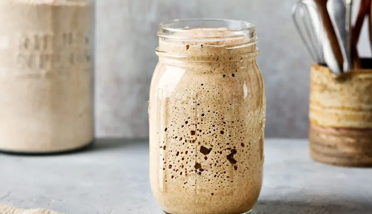 Aktiver Sauerteig im Glas, bereit für das Ansetzen mit dem Thermomix. Daneben ein Glas mit Mehl und ein Behälter mit Küchenutensilien.