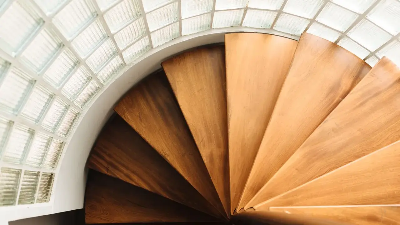 Escalera helicoidal de madera con peldaños que giran bajo un techo de bloques de vidrio translúcido.