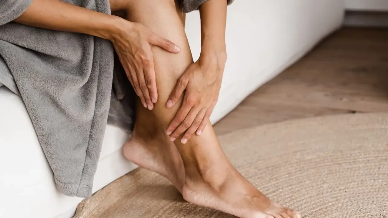 Mani che accarezzano le gambe, evidenziando la pelle secca. La persona è seduta su un letto, avvolta in un accappatoio grigio.