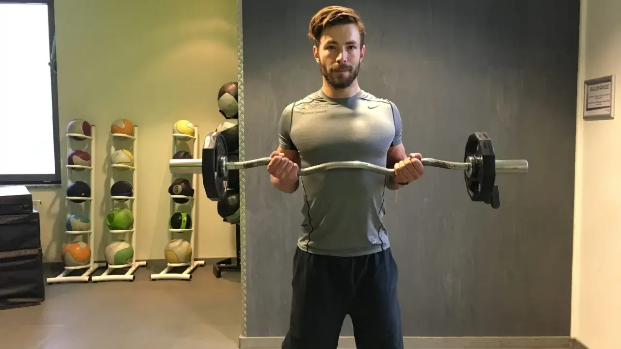 Mann macht Bizeps-Curls mit einer Langhantel. Im Hintergrund stehen Medizinbälle.