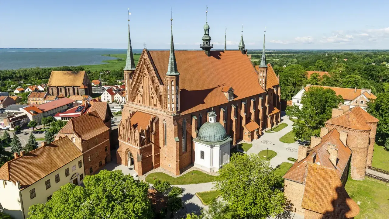 Zamek Frombork z katedrą i wieżami, otoczony zielenią, nad błękitną wodą.