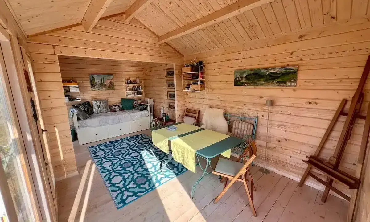 Gemütlicher Gartenhaus Innenausbau mit Tagesbett, Esstisch und Staffelei. Holzwände und Decke schaffen eine warme Atmosphäre.