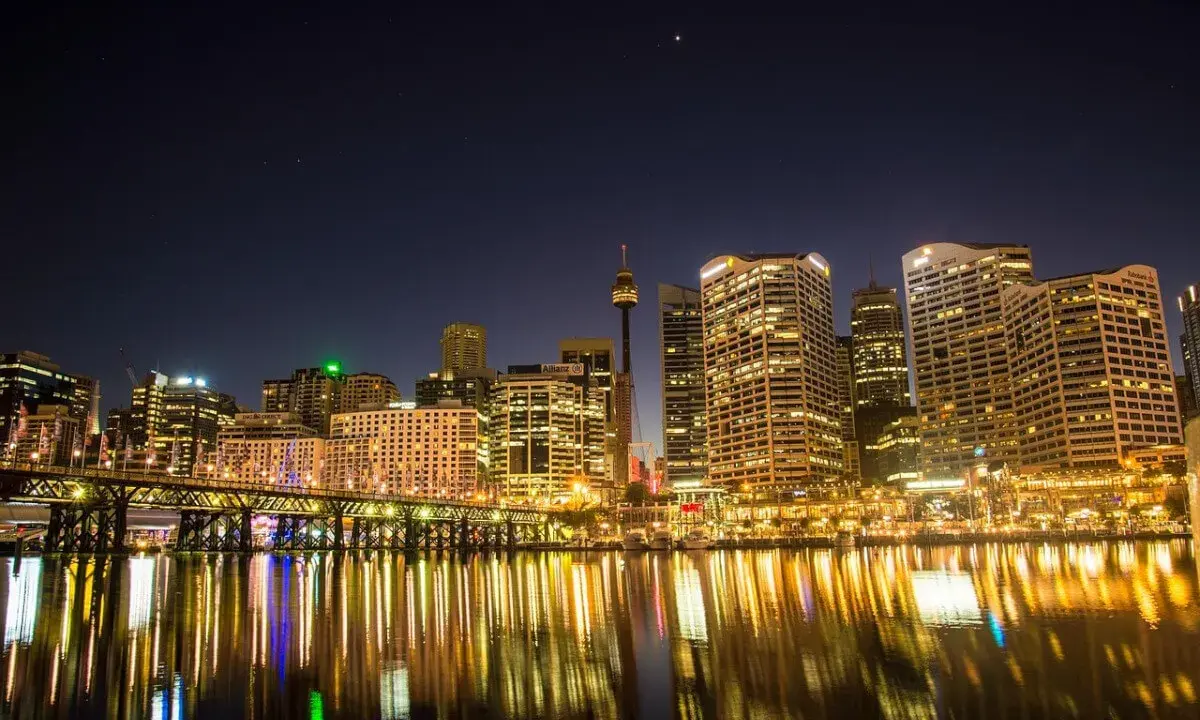 Sydney nocą, z mostem i odbijającymi się w wodzie światłami. Zastanawiasz się, ile się leci do Australii, patrząc na ten widok?