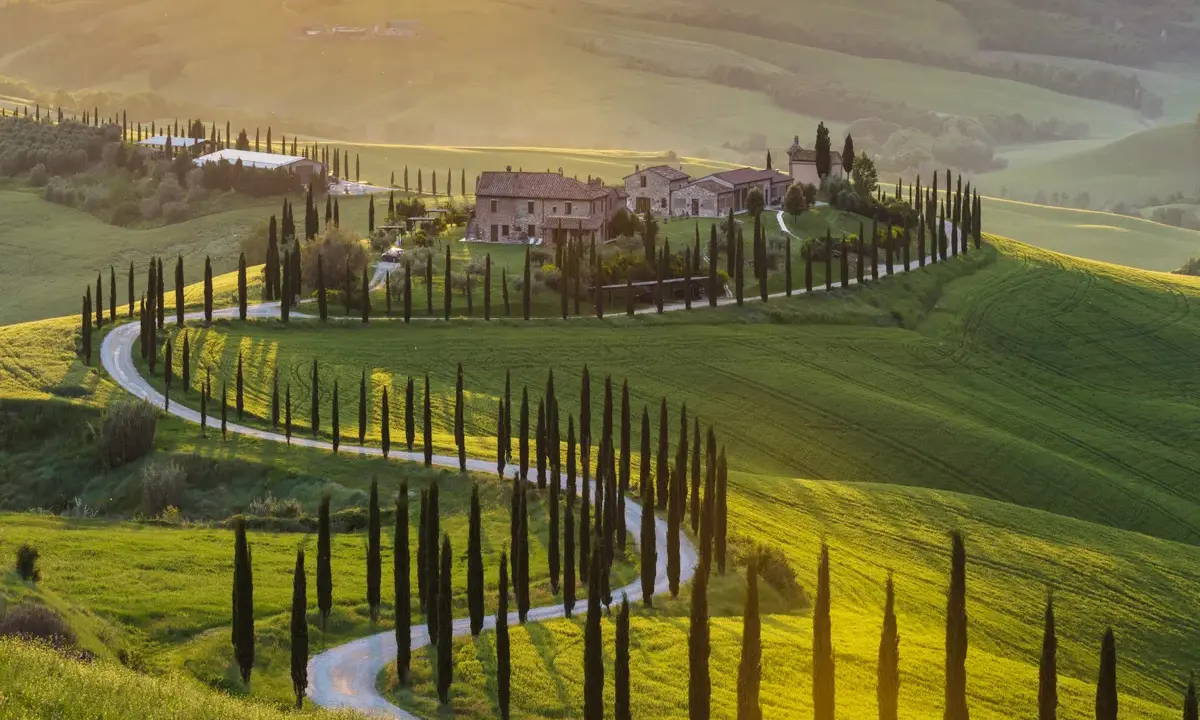 Colline toscane al tramonto, cipressi lungo una strada tortuosa, un casale in pietra. Ecco cosa vedere in Val d'Orcia.